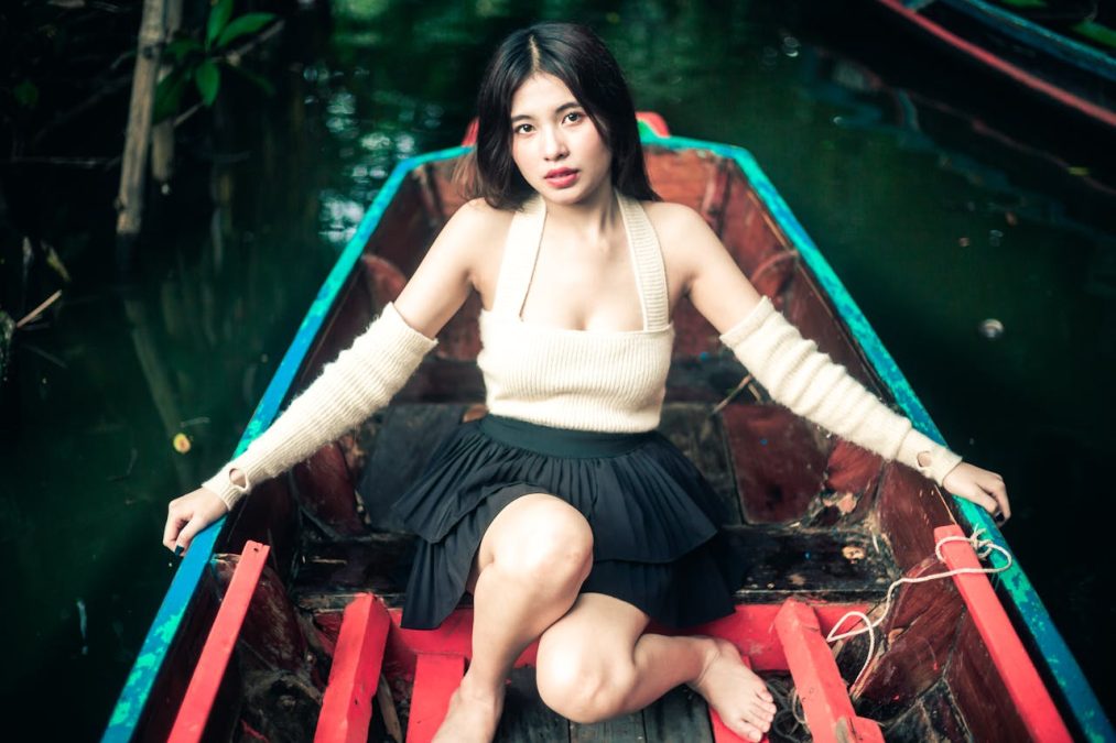 A woman sitting in a colorful wooden boat, wearing a cream sweater and black skirt, surrounded by water and greenery.