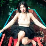 A woman sitting in a colorful wooden boat, wearing a cream sweater and black skirt, surrounded by water and greenery.