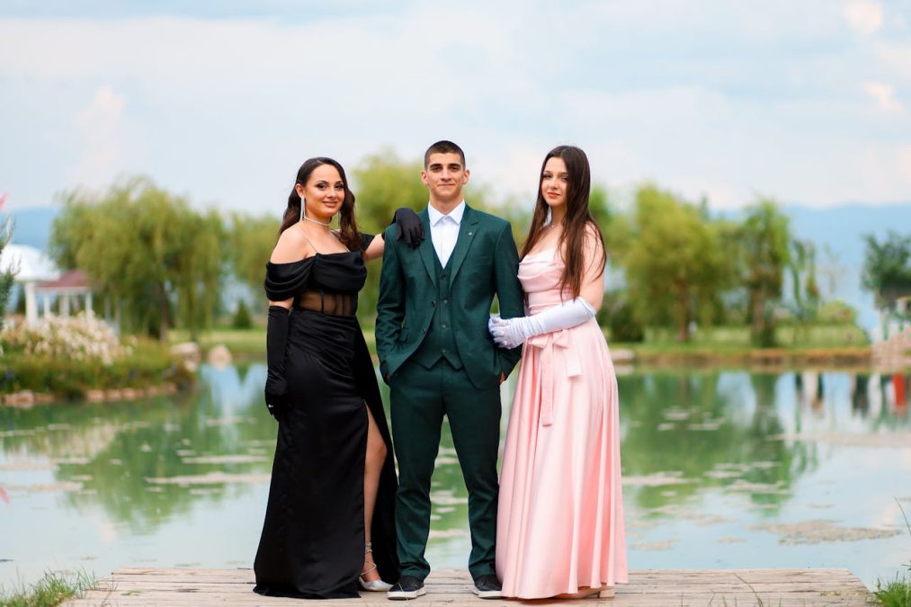 Group of three individuals in formal attire posing by a serene water body, showcasing elegant dresses and a suit, perfect for event photography.